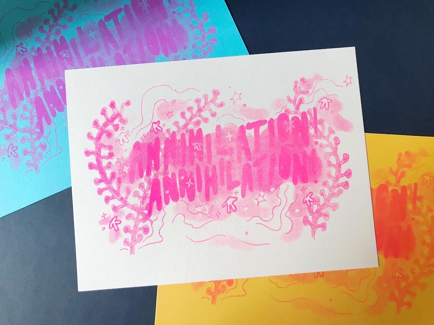 Three risograph prints of the same design, the words 'ANNIHILATION! ANNIHILATION! surrounded by plants, clouds, stars and spores, all coloured in halftone. The designs have been printed in pink on white, red on orange and purle on blue.