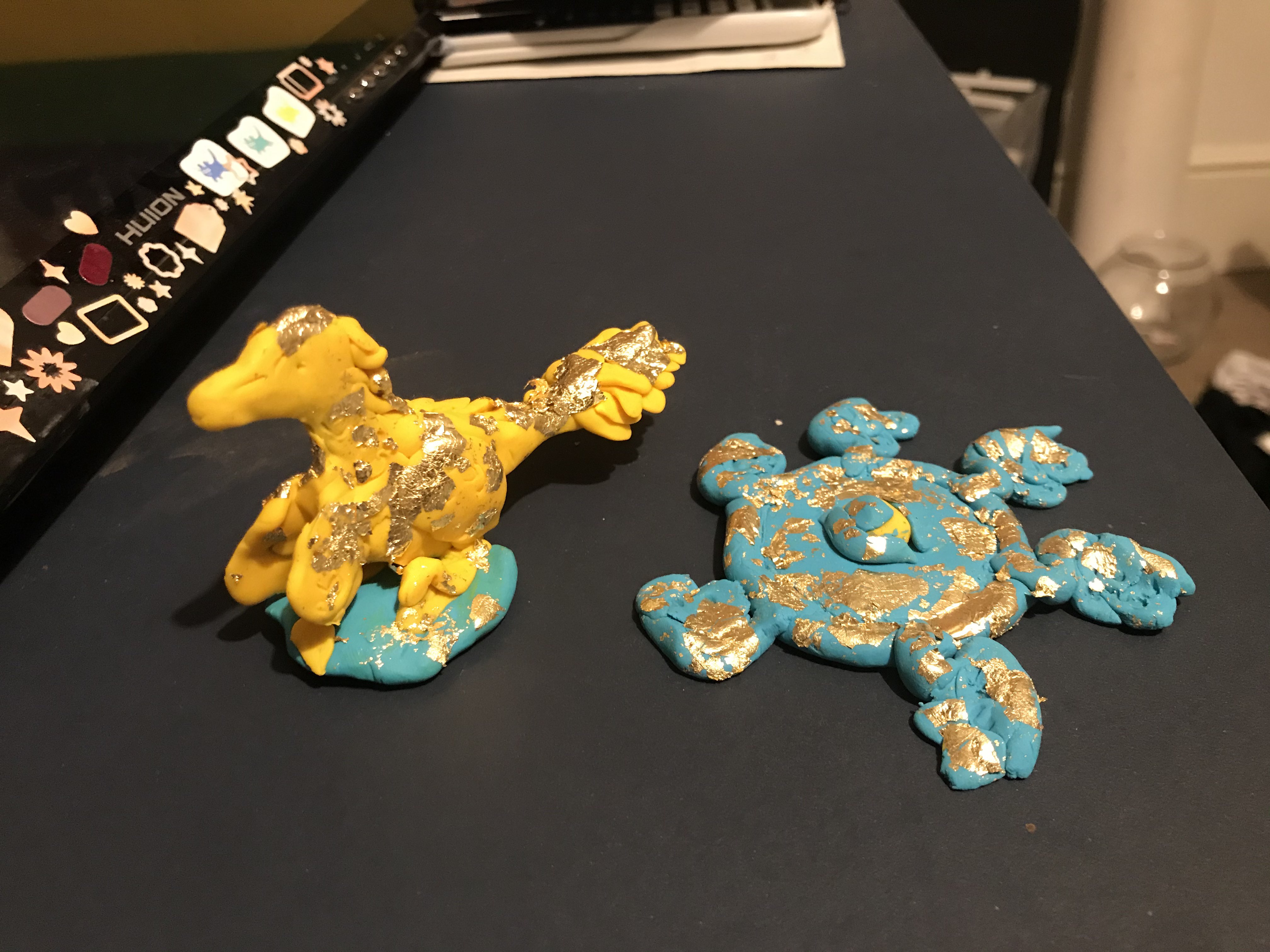 a yellow fimo velociraptor dinosaur and a blue fimo angel, both with gold flakes on