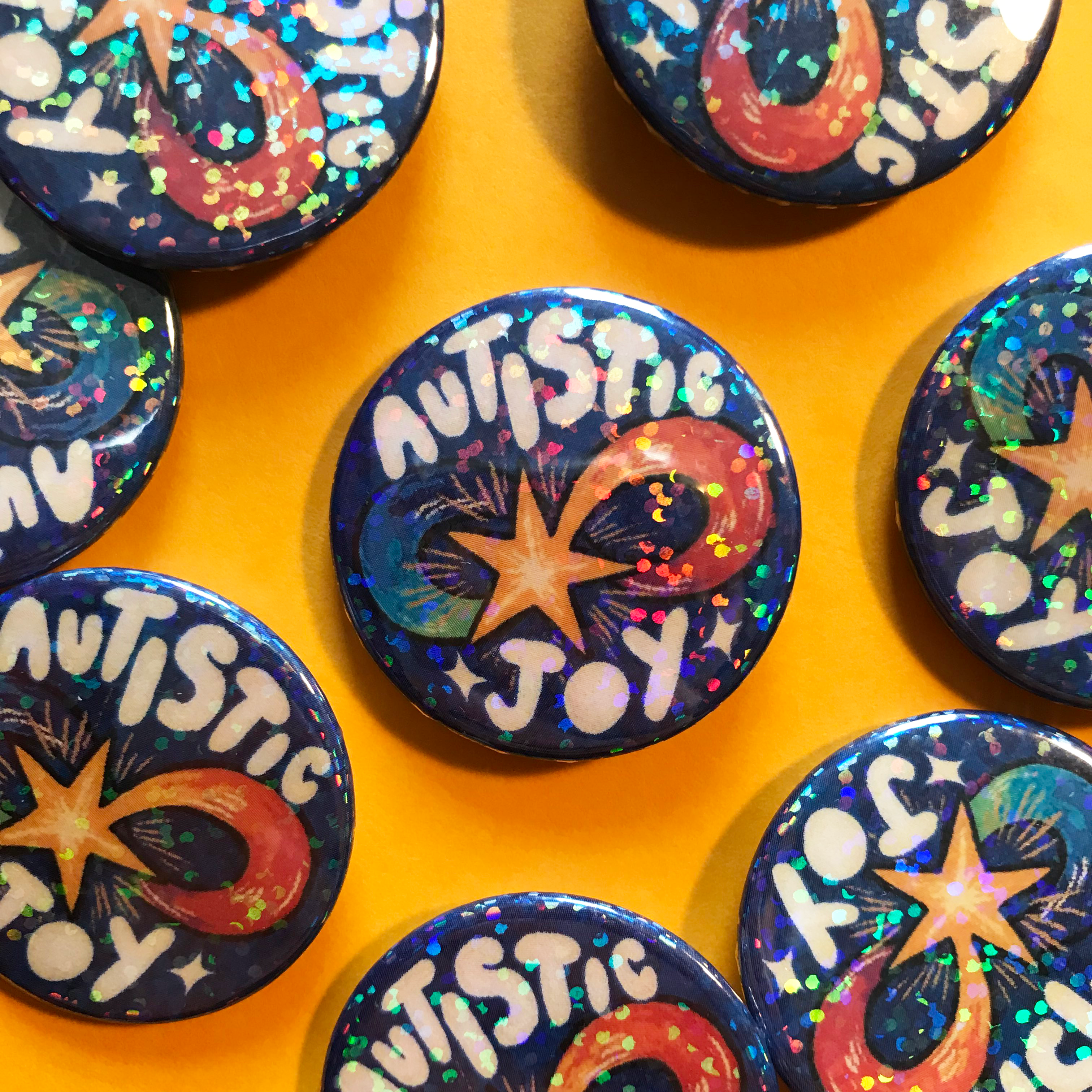 A photo of holographic badges against a yellow background. The badges have a design of a rainbow covered infinity sign shaped shooting star against a dark blue background, with text that says 'autistic joy'