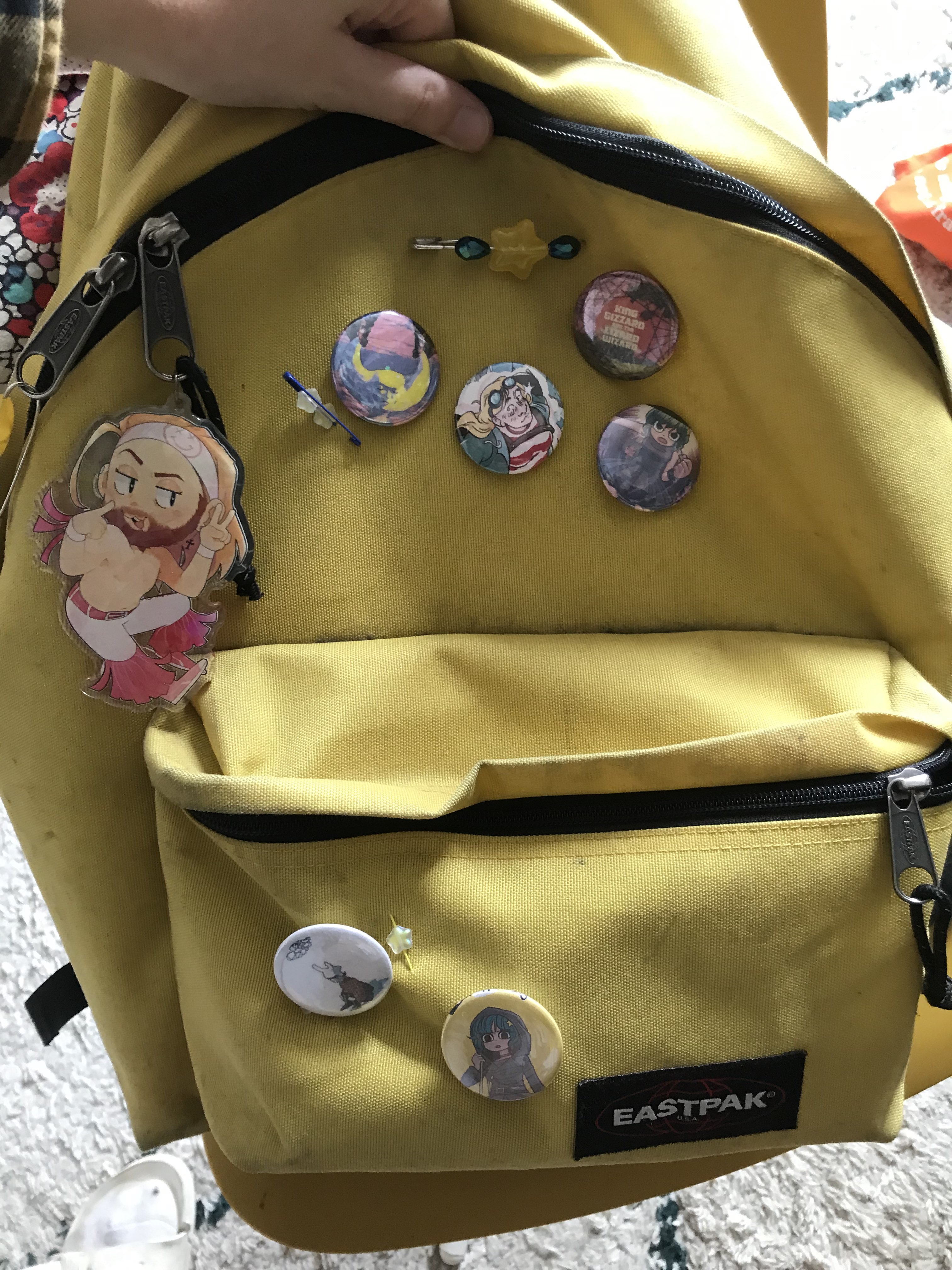 A photo of a yellow backpack with multiple round button badges on, with album covers, video game characters and comic panels on, some with holographic overlay on.