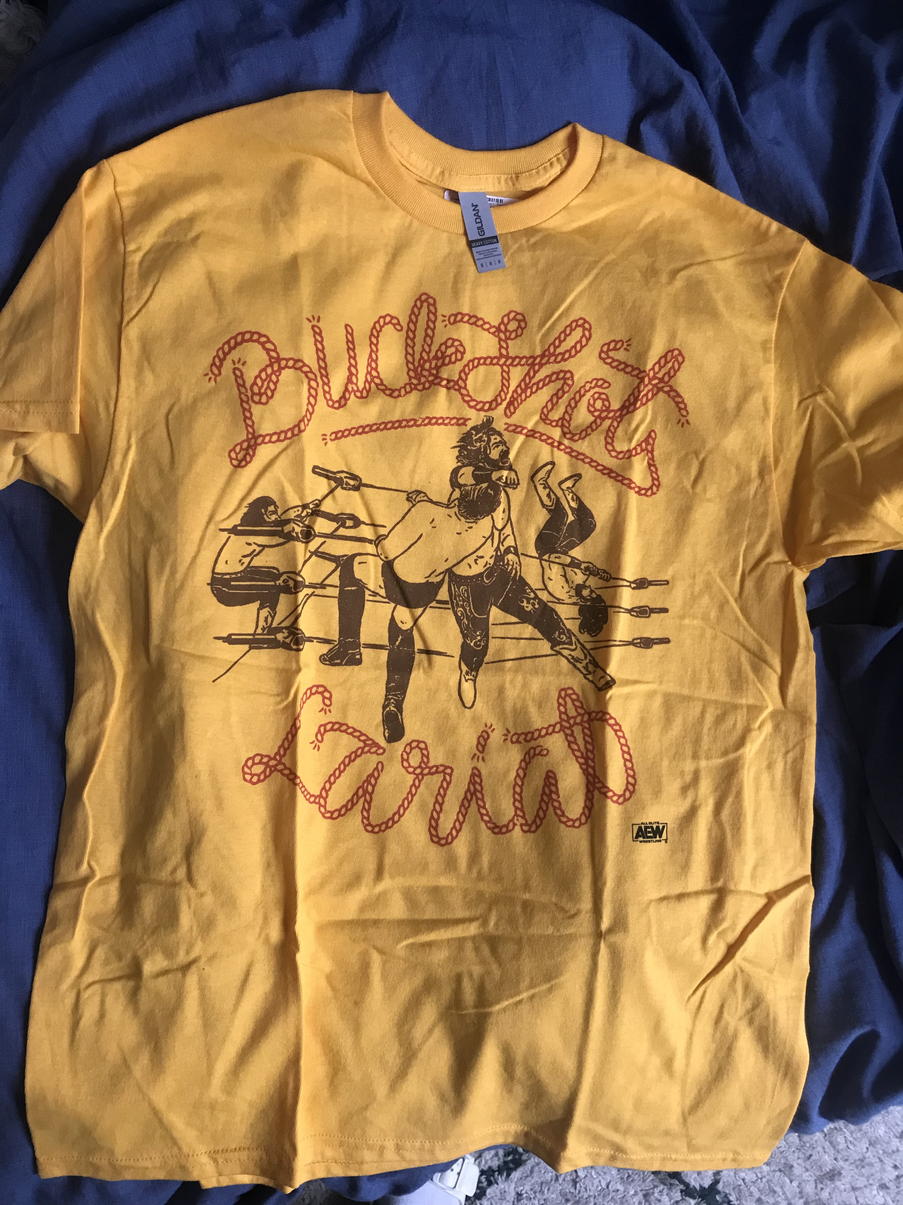 A yellow tshirt with a one-colour illustration in black of a pro wrestler throwing a lariat, and with text surrounding it in red with a stylized rope effect that says Buckshot Lariat.