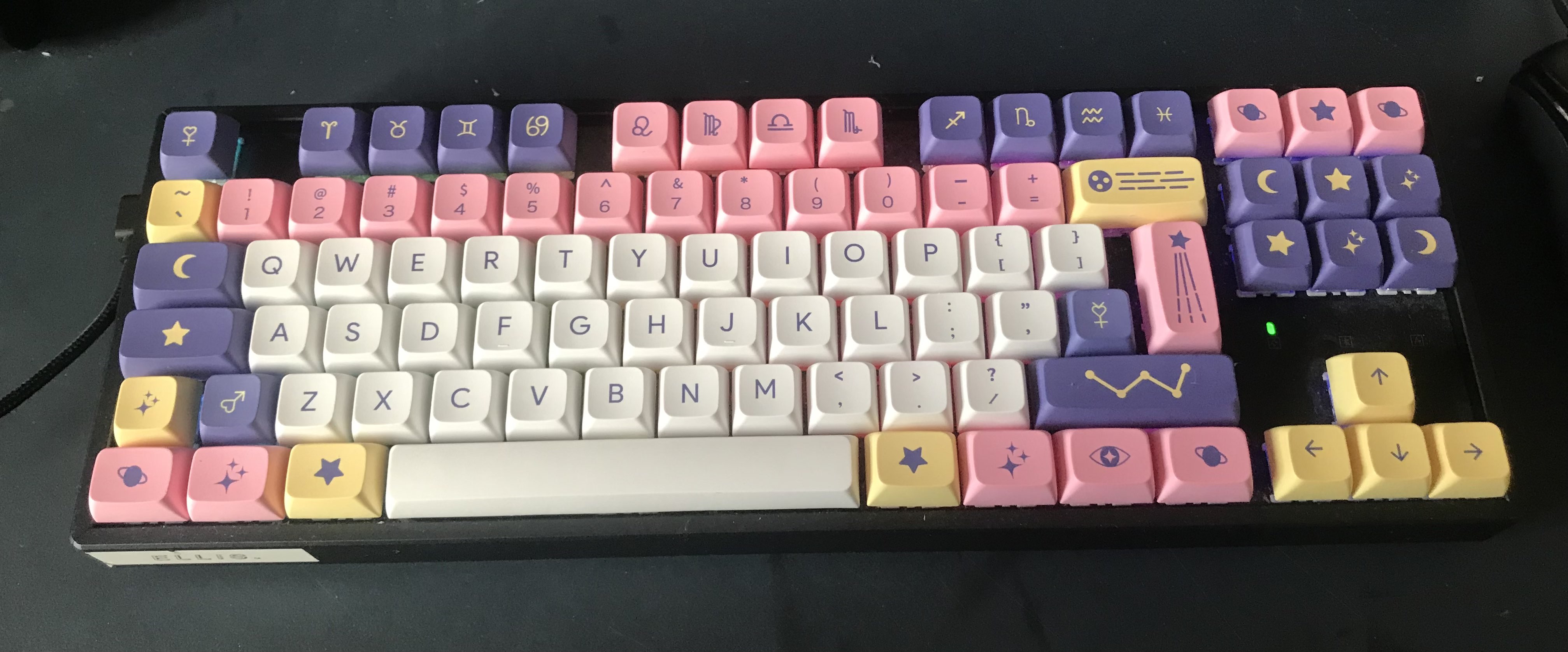A photo of a mechanical keyboard with keycaps in white, pink, yellow and purple colours, with star and astrology decals on the special keys.