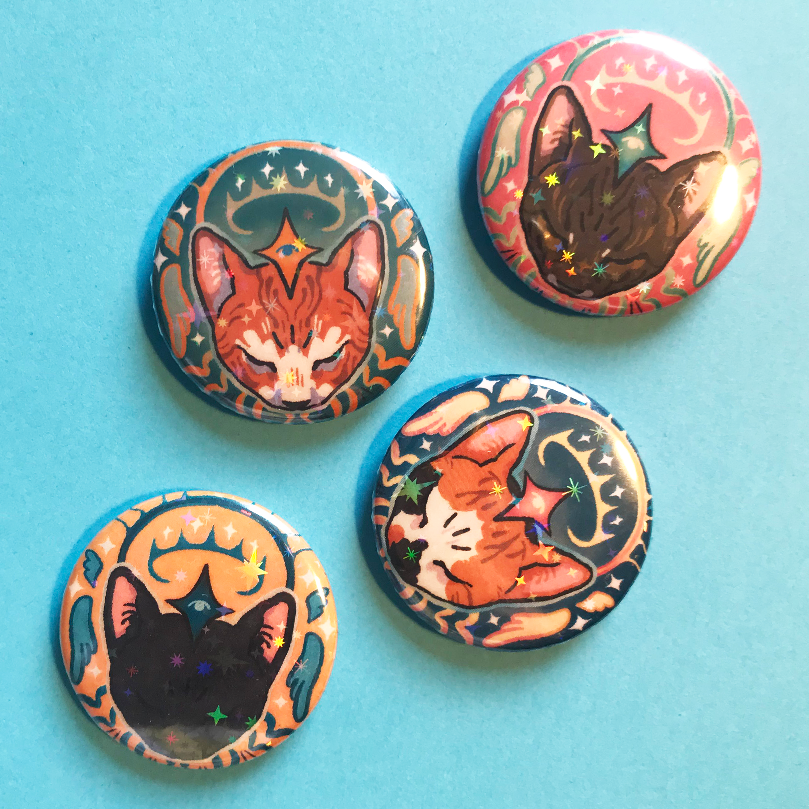 A photo of four badges on a blue background. The design on each is of an illustrated cats head surrounded by drawn halo and wing-esque motifs. Each badge has a different colour scheme and fur pattern of cat.