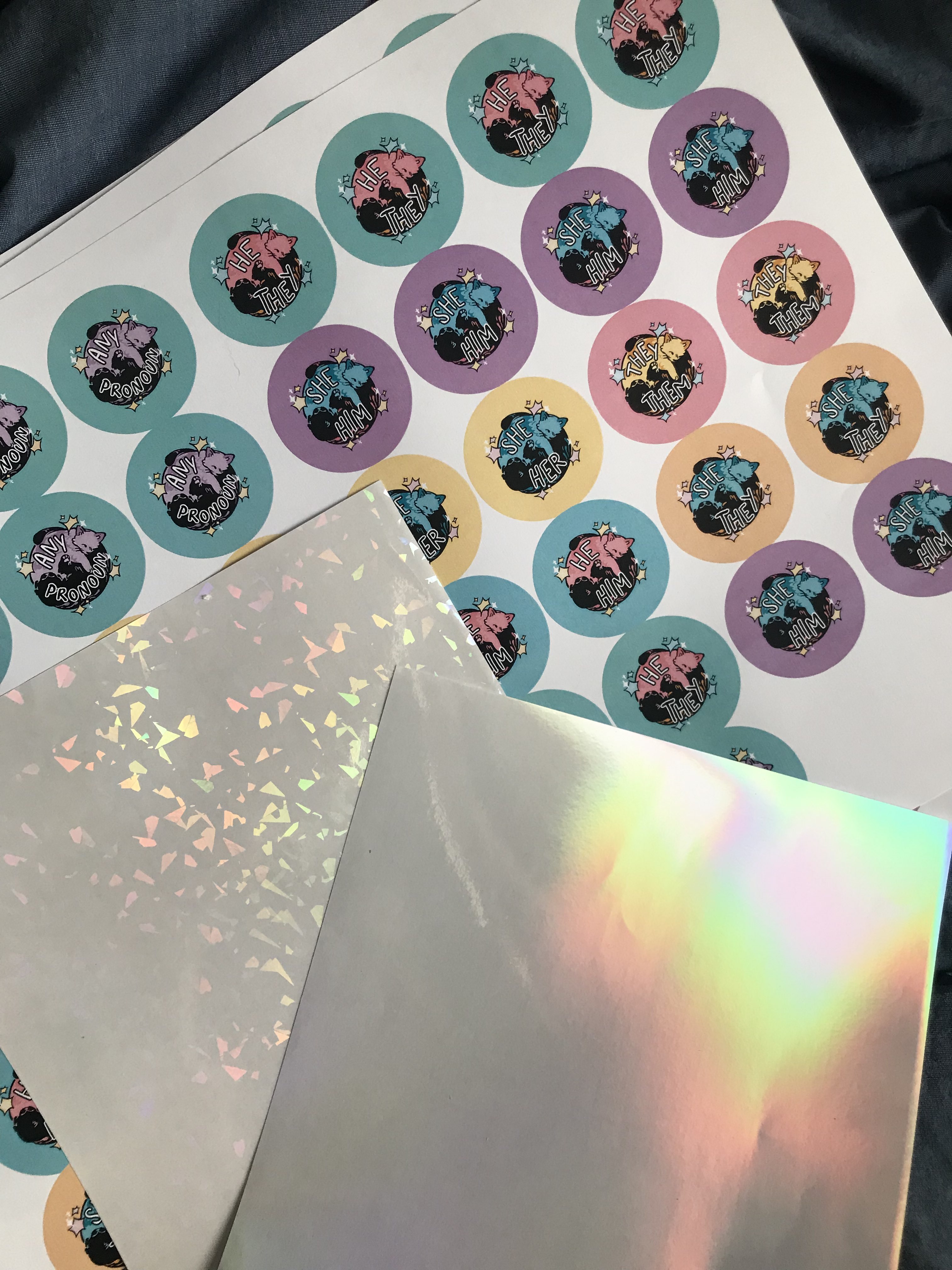 A photo of a piece of paper covered in printouts of a digital drawing ready to be turned into badges of cat-themed pronoun badges. There are also two holographic sheets with different patterns.