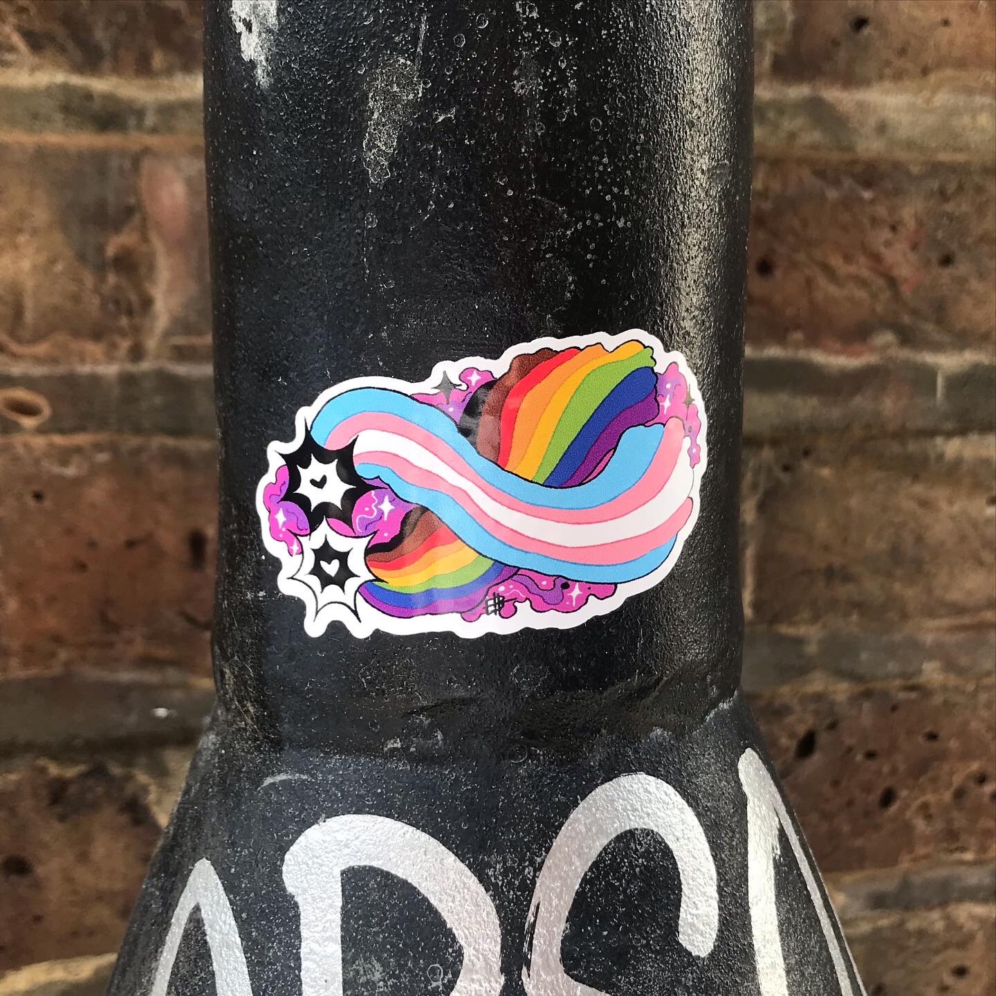 A sticker of Two shooting stars, each with a heart at the centre, one black, one white, with  comet trails of the trans and philadelphia rainbow flag respectively, surrounded by a purple nebula, wrapped around a lamppost.