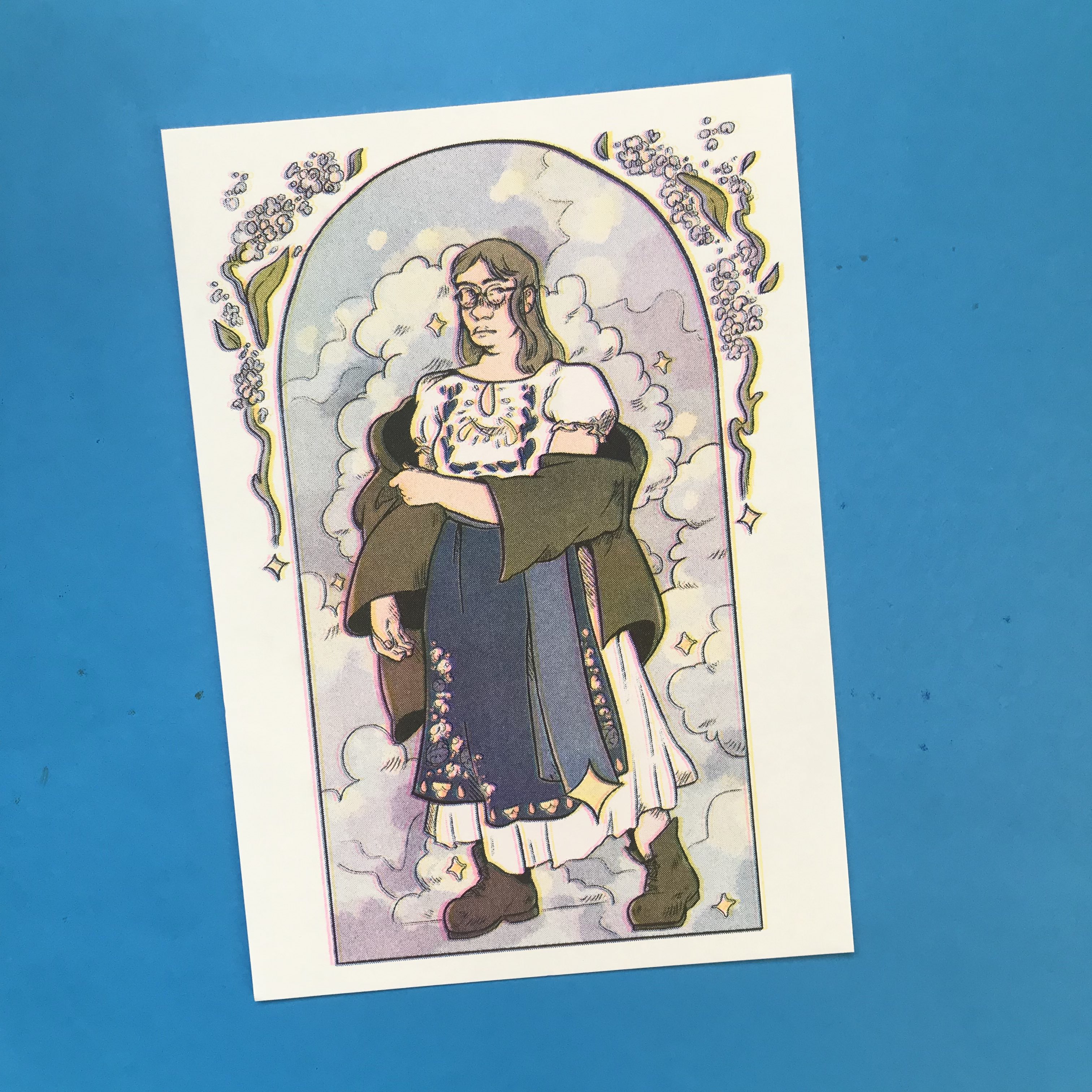 A photo of a full-colour CMYK risoprint against a blue background. The print is an illustration of a young White woman in traditional Eastern European-inspired folk dress in blue and an oversized brown jacket. Behind her is a arch frame full of blue clouds, with details of stars and flowers on the edge of the frame.