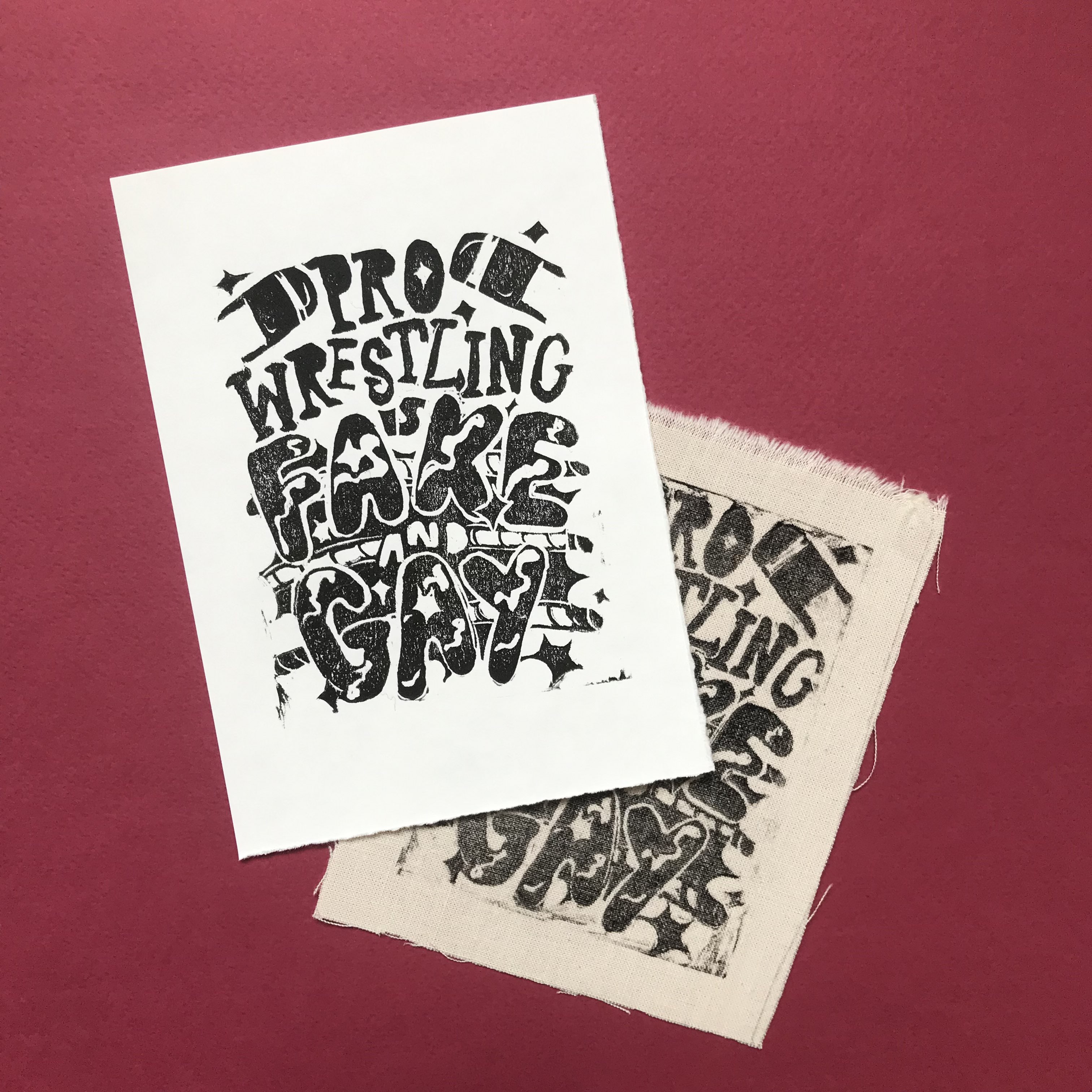A product photo against a dark red background of black linoprint in black on white paper and fabric of stylized bubble text reading 'PRO WRESTLING IS FAKE AND GAY' with ring ropes reminiscent of a wrestling ring behind it.